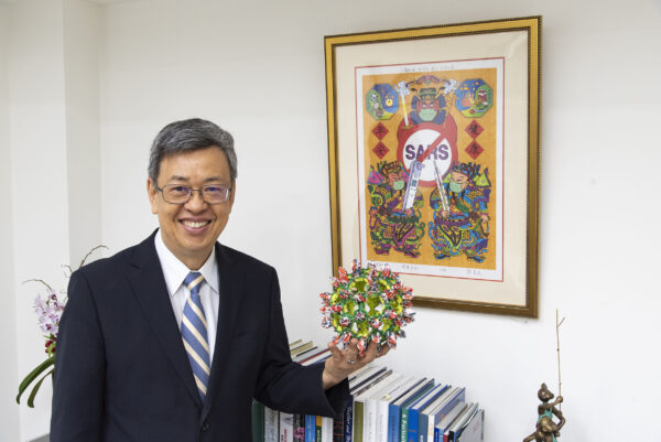 陳建仁院士手拿冠狀病毒摺紙,身後是藝術家謝省民為紀念 SARS抗疫而創作的版畫〈健康平安〉圖|陳建仁